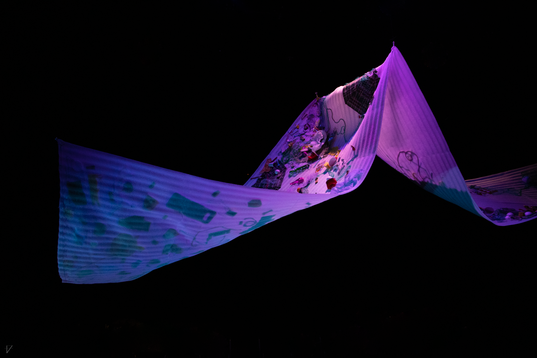 Drowning Voices set piece, a large swooping sheet of flexible styrofoam with garbage stuck to it, lit in purple and blue, from a distance