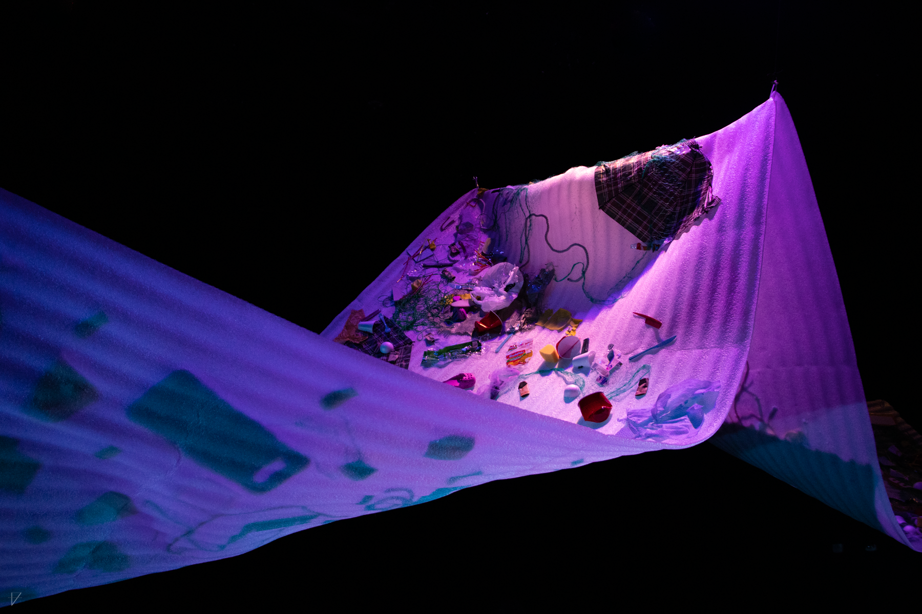 Drowning Voices set piece, a large swooping sheet of flexible styrofoam with garbage stuck to it, lit in purple and blue, from a medium distance