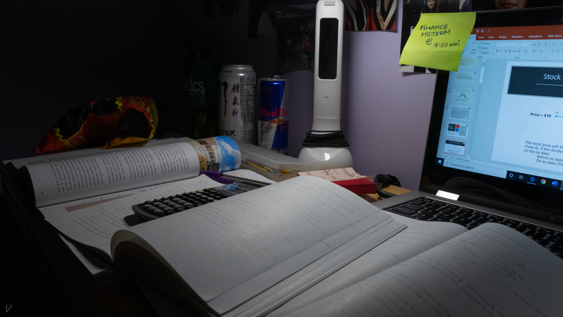 A student's desk at night, covered in textbooks and snacks, with a sticky note that reads Finance Midterm at 9 AM