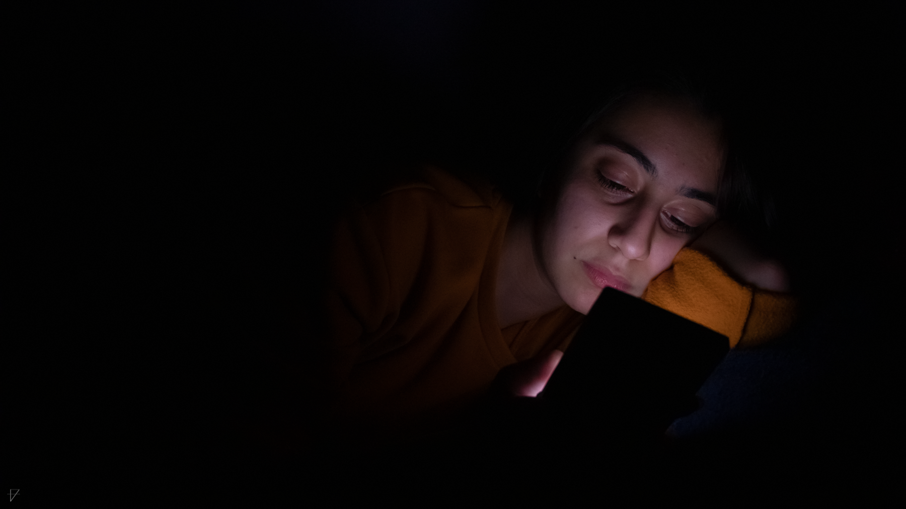 Close up of a girl in the dark, her face lit only by her phone as she lies in bed